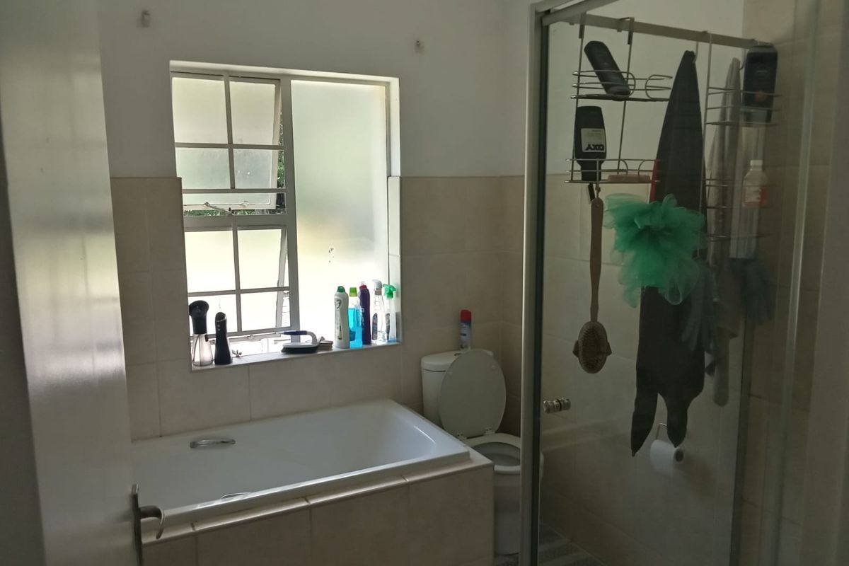 A functional bathroom featuring a built-in bathtub, a separate glass-enclosed shower, and a toilet, with neutral wall tiling and a window for natural light.