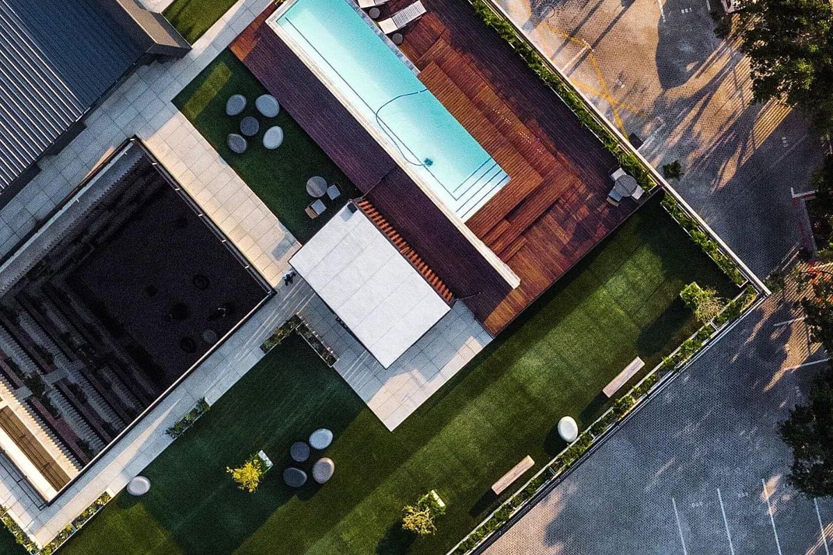 Aerial view of a modern apartment complex featuring a swimming pool with timber decking, landscaped green spaces, and paved parking areas.
