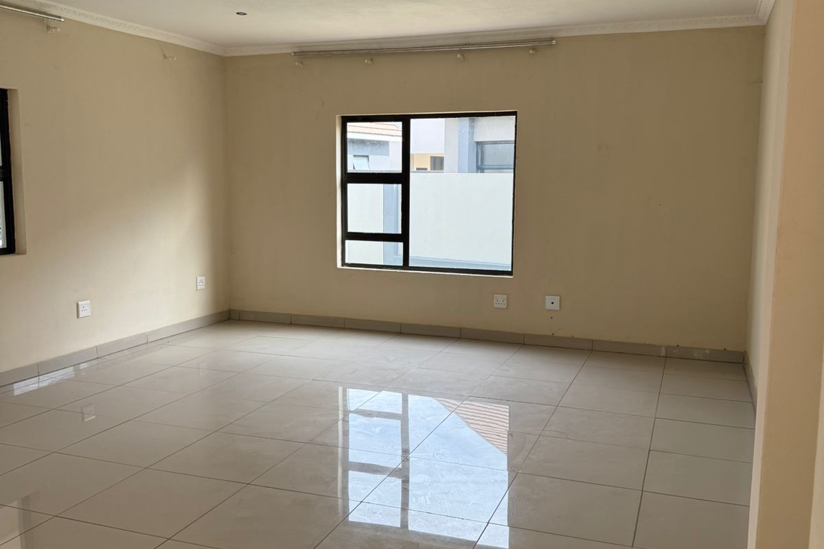 A spacious, empty living area featuring neutral tiled flooring, recessed ceiling lighting, and large windows providing natural light.