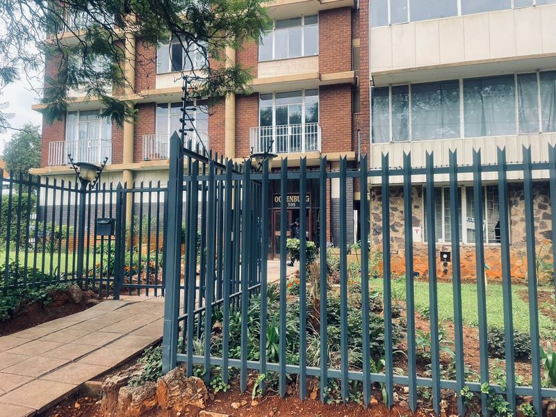 Front elevation of the Ocenburg apartment complex, featuring a brick facade, secure perimeter fencing, and a paved pedestrian entrance path.