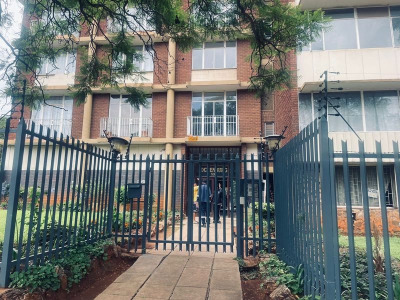 Front exterior view of a multi-story brick apartment building featuring secure metal perimeter fencing, gated pedestrian access, and balconies.
