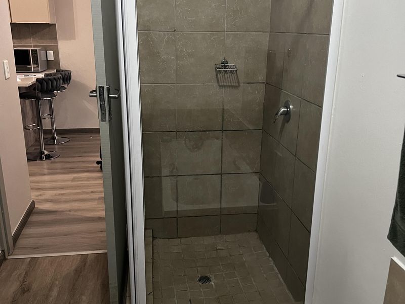 A bathroom featuring a tiled shower enclosure with a glass door, adjacent to a view of the kitchen area.