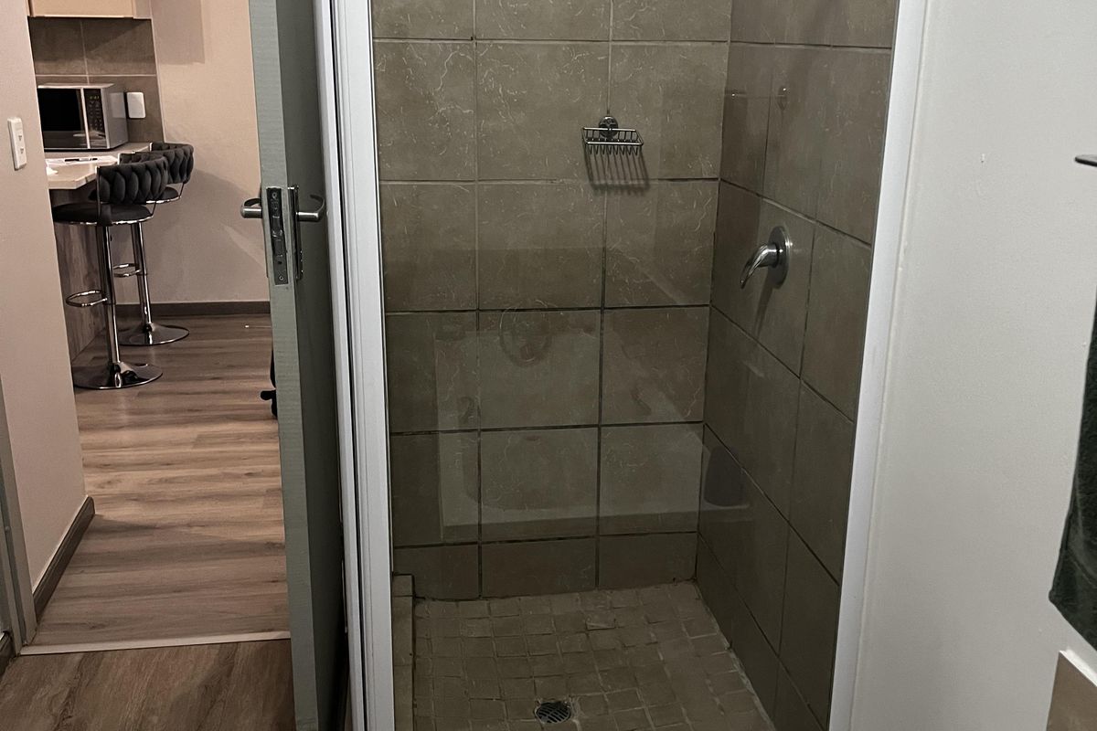 A bathroom featuring a tiled shower enclosure with a glass door, adjacent to a view of the kitchen area.