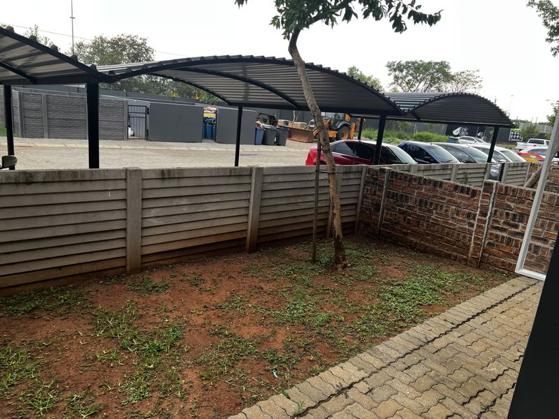 A small outdoor garden area featuring a paved walkway, a concrete boundary wall, and a covered parking structure in the background.