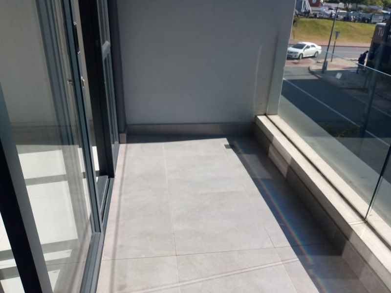 A modern apartment balcony featuring grey floor tiles, a glass balustrade, and sliding door access, offering a view of the street below.