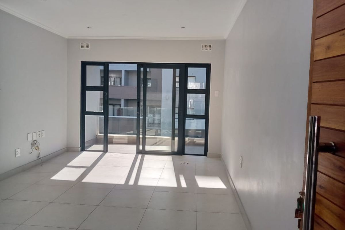 A bright, empty living area featuring large sliding glass doors that open onto a balcony, with neutral tiled flooring and recessed ceiling lighting.