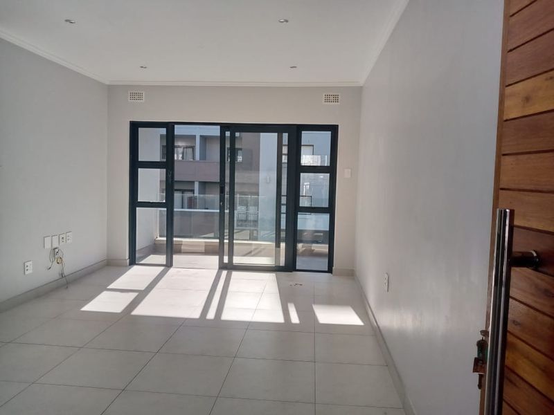 A bright, empty living area featuring large sliding glass doors that open onto a balcony, with neutral tiled flooring and recessed ceiling lighting.