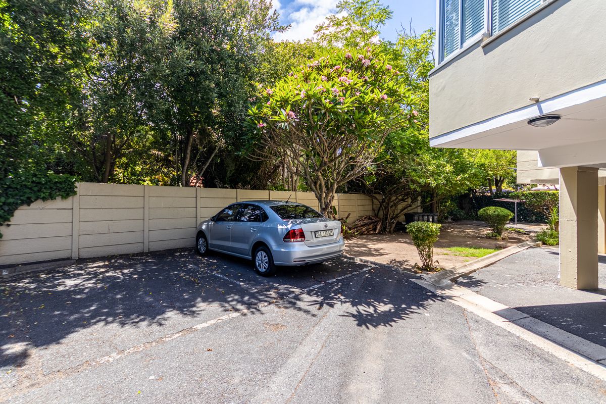 Exterior view of the apartment complex parking area featuring paved bays, mature trees, and a boundary wall.