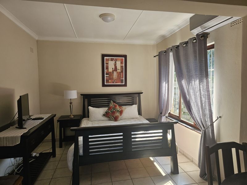 A well-lit bedroom featuring tiled flooring, an integrated air conditioning unit, and a window fitted with security bars.