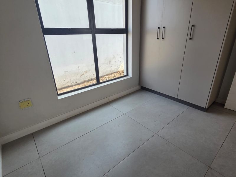 A bright, empty bedroom featuring modern large-format floor tiles, built-in cupboards, and a large aluminium-framed window.