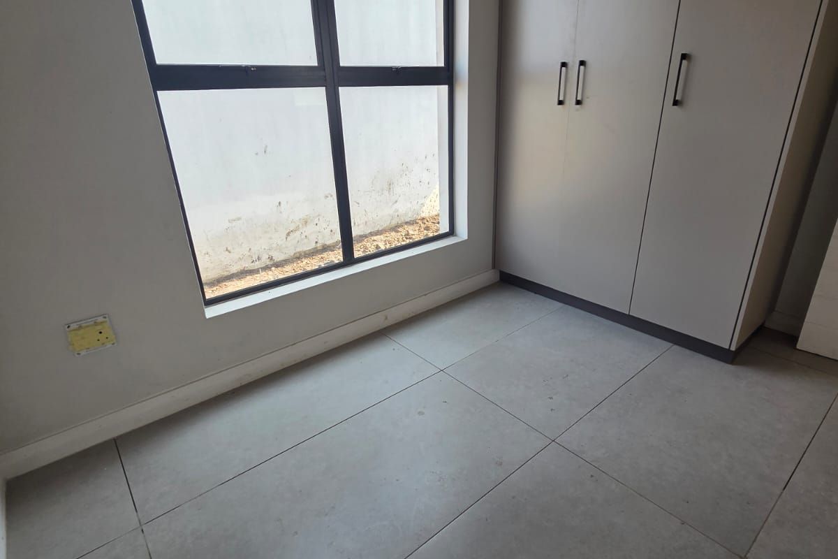 A bright, empty bedroom featuring modern large-format floor tiles, built-in cupboards, and a large aluminium-framed window.