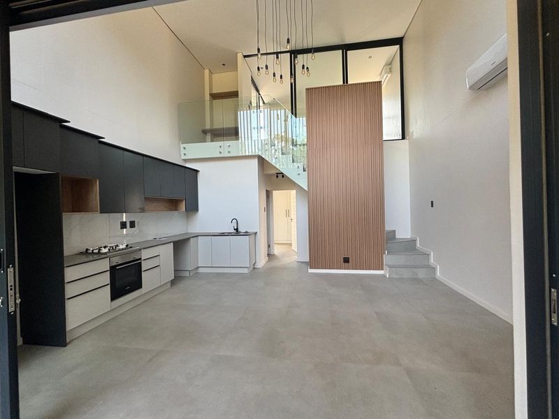 Modern open-plan living area featuring a sleek kitchen with built-in appliances, double-volume ceilings, a mezzanine level with glass balustrades, and contemporary timber feature wall.