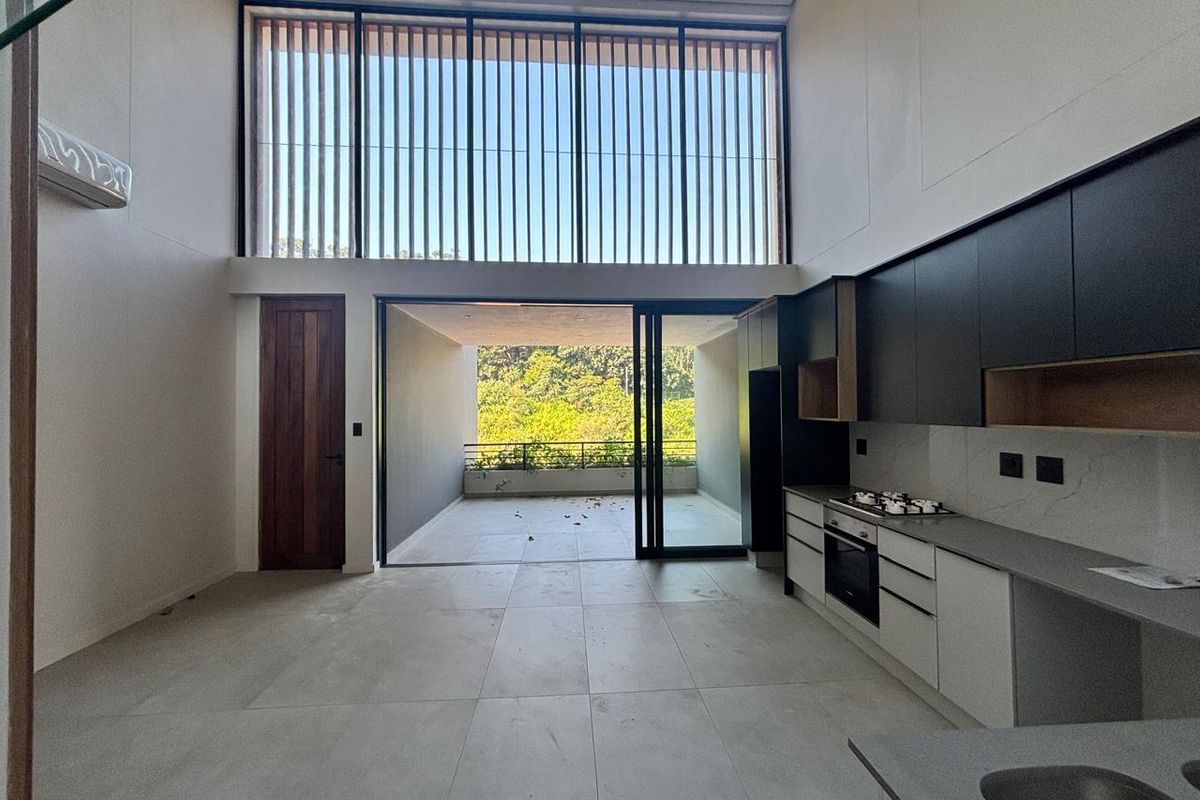 Modern open-plan kitchen and living area featuring double-height ceilings, sleek cabinetry, integrated appliances, and seamless flow to a covered patio with garden views.