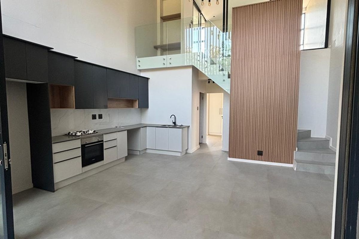 Modern open-plan kitchen and living area featuring high ceilings, sleek cabinetry, integrated appliances, a timber feature wall, and a contemporary glass-railed staircase.