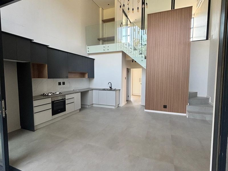 Modern open-plan kitchen and living area featuring high ceilings, sleek cabinetry, integrated appliances, a timber feature wall, and a contemporary glass-railed staircase.