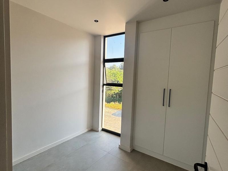 A bright, modern, and empty bedroom featuring neutral tiled flooring, recessed downlights, and a built-in wardrobe with clean white finishes.