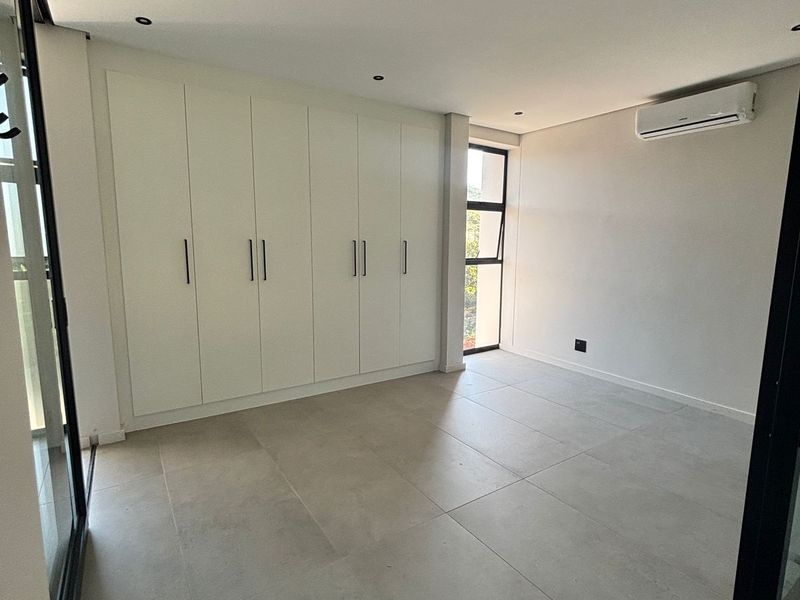 A bright, modern bedroom featuring neutral tiled flooring, extensive built-in cabinetry, and a wall-mounted air conditioning unit.