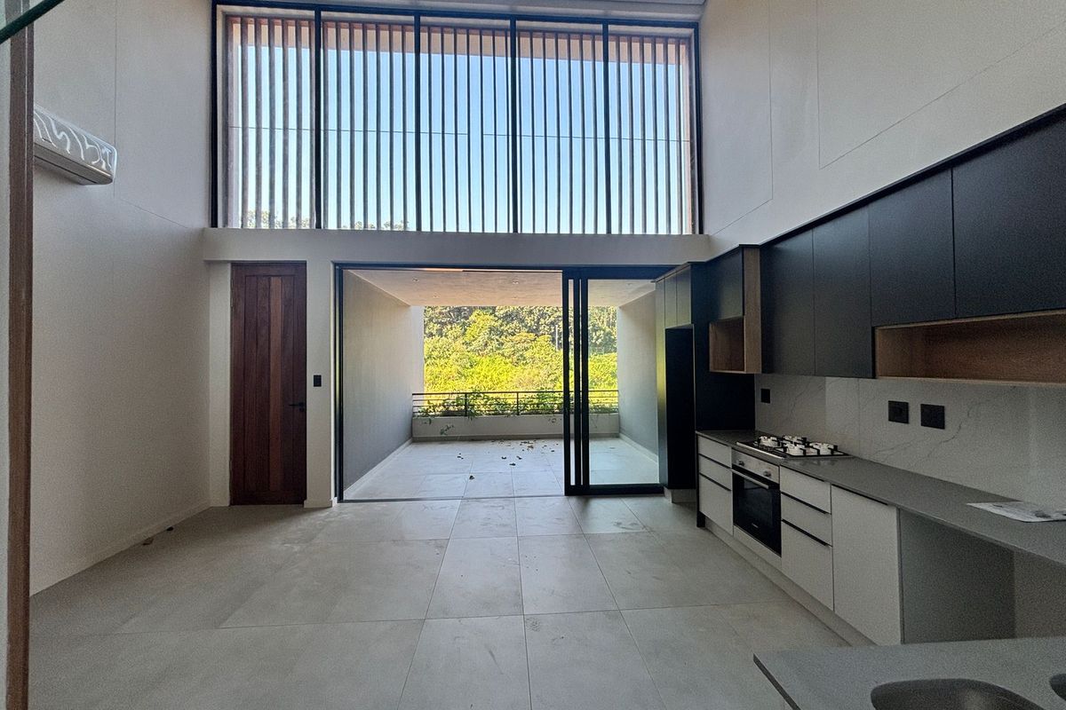 Modern open-plan kitchen and living area featuring double-height ceilings, sleek cabinetry, integrated appliances, and seamless flow to a covered patio with garden views.