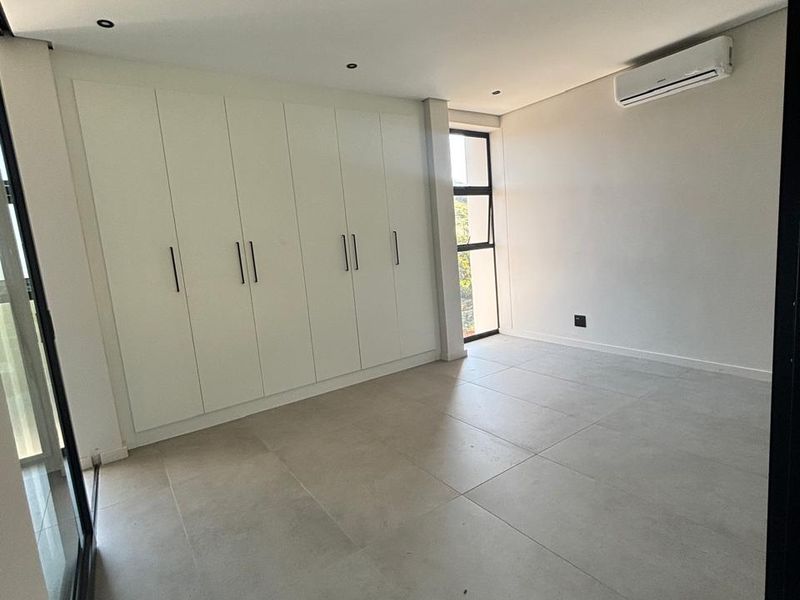 A bright, modern, and empty bedroom featuring large built-in cupboards, tiled flooring, and a wall-mounted air conditioning unit.