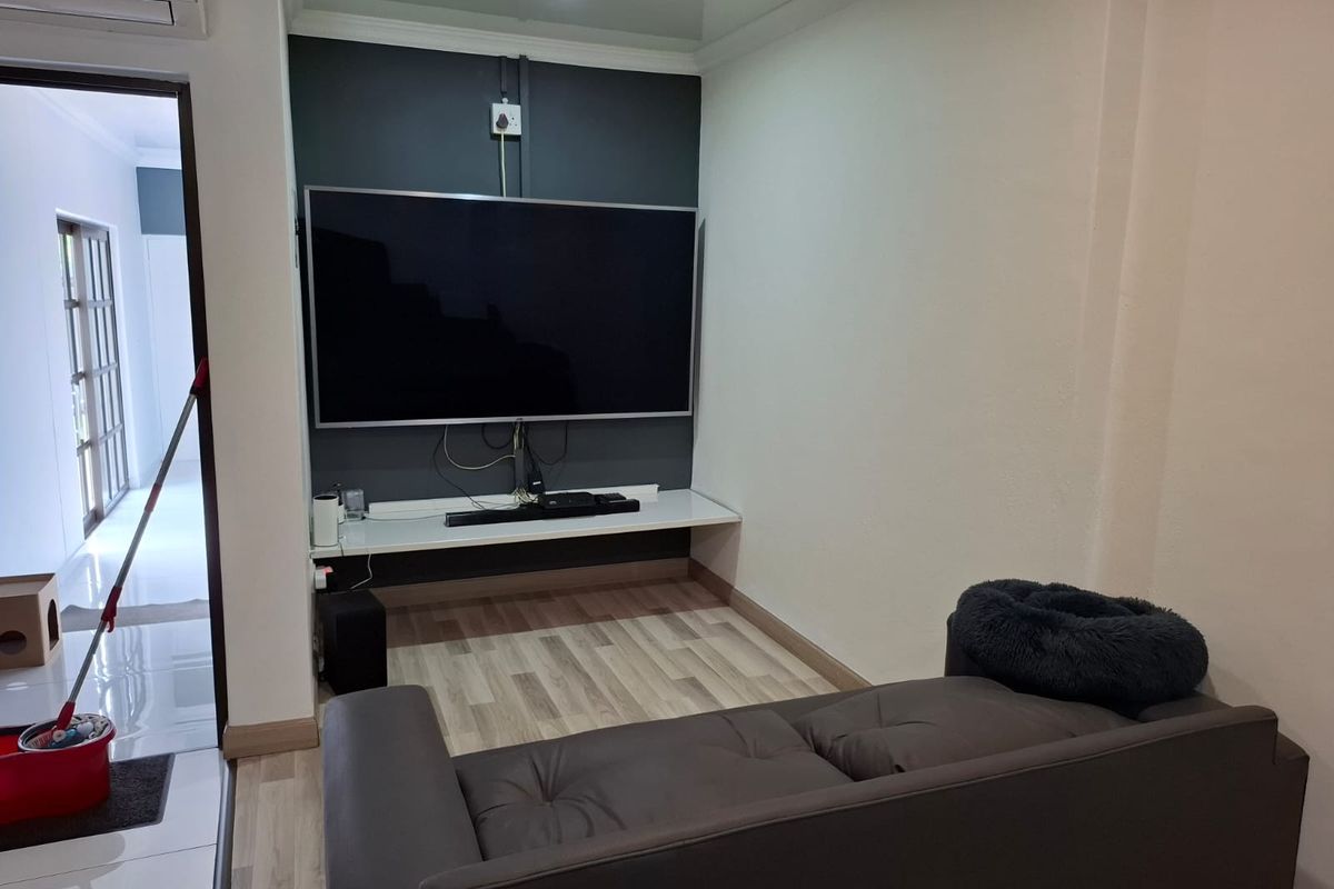 A living room featuring a wall-mounted television, floating shelf, and light-toned laminate flooring, with an adjacent hallway visible.