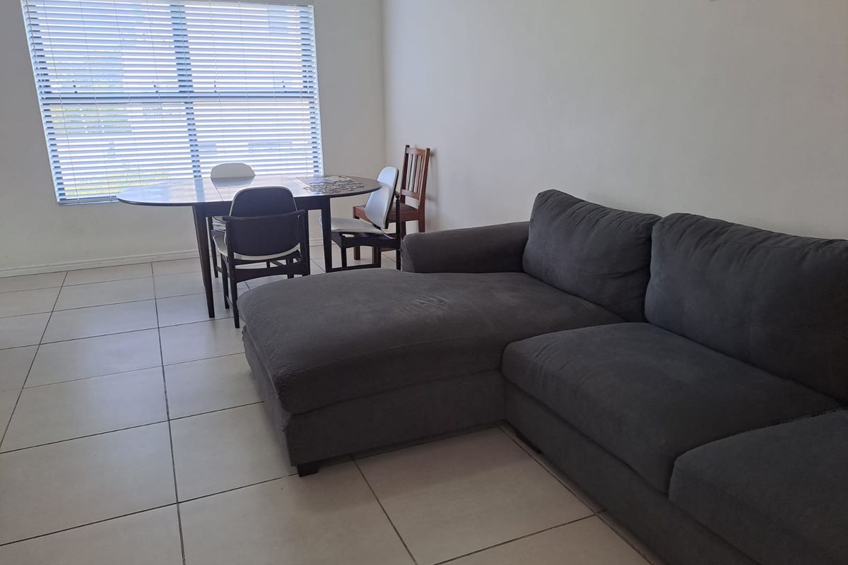 A living area featuring neutral tiled flooring and large windows fitted with venetian blinds, providing a bright and functional space.
