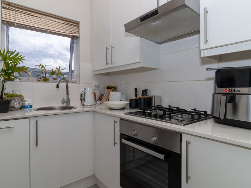 A modern, compact kitchen featuring white cabinetry, stone countertops, a gas hob, built-in oven, and a stainless steel sink with a tiled splashback.