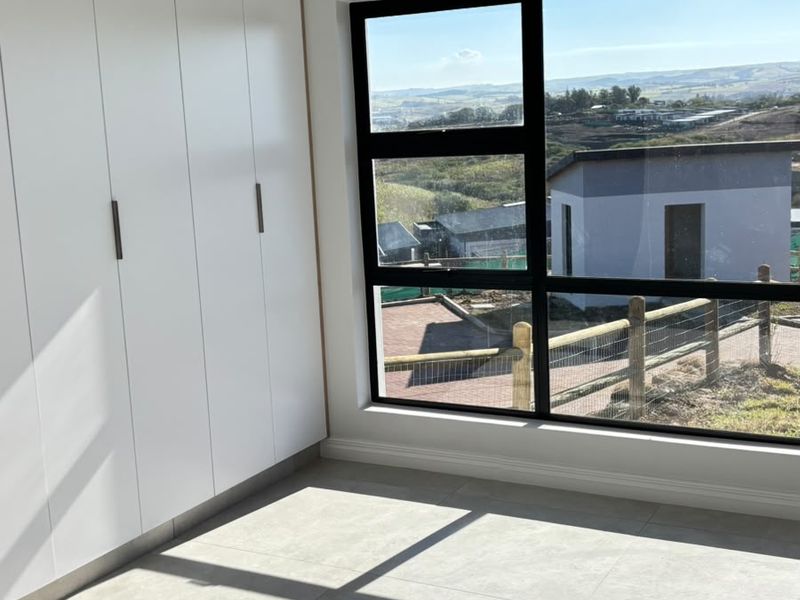 A bright, modern bedroom featuring built-in cupboards, large format floor tiles, and a large window offering scenic views.