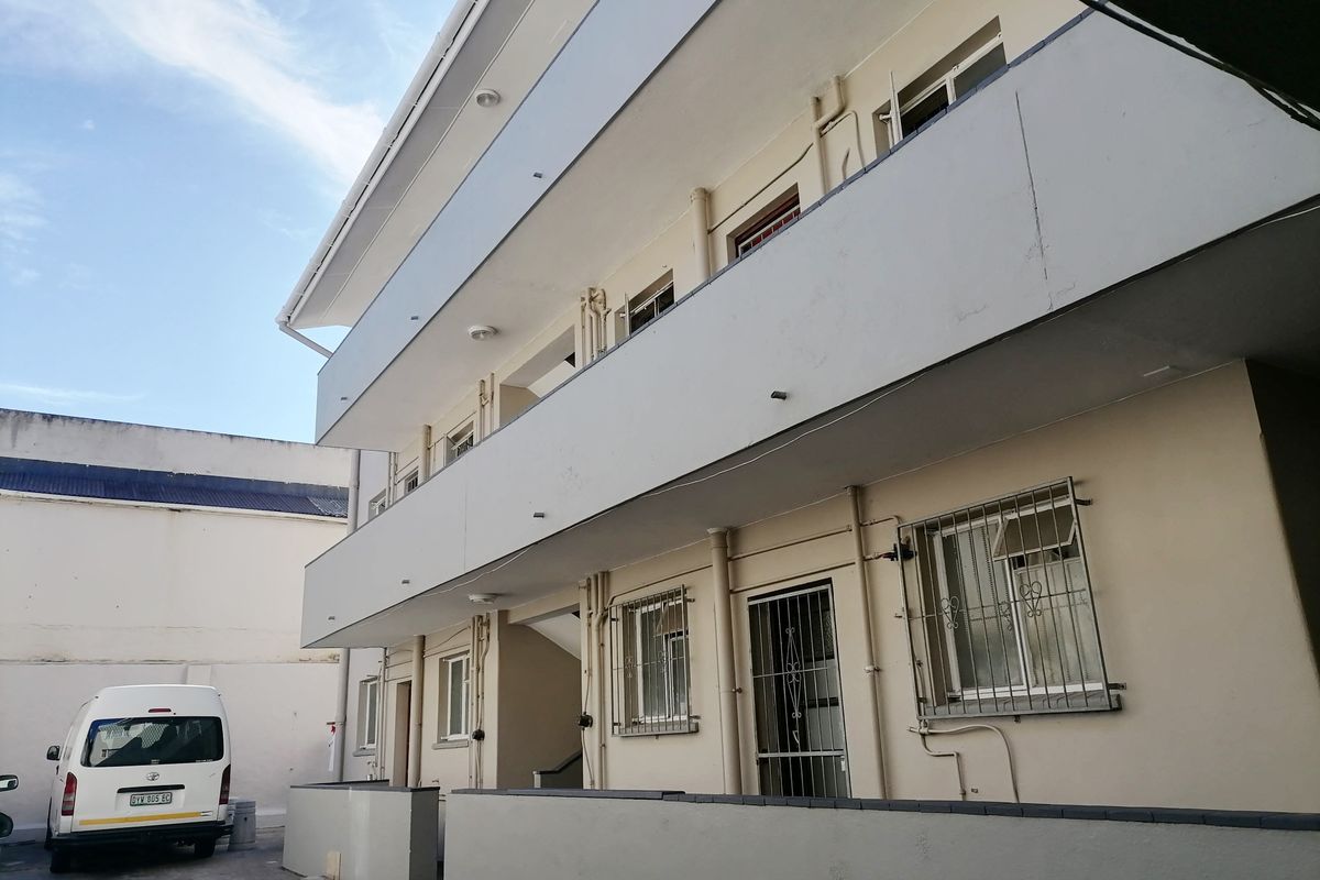 Exterior view of a multi-story apartment building featuring balconies, security bars on windows, and a paved ground level area.