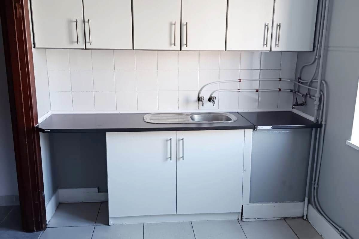 A functional kitchen area featuring white cabinetry, a stainless steel sink, tiled backsplash, and tiled flooring with exposed plumbing fixtures.