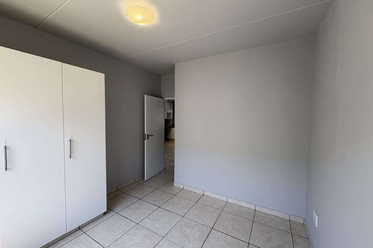 A bedroom featuring neutral grey walls, tiled flooring, and a built-in white wardrobe, with a doorway leading to an adjacent room.