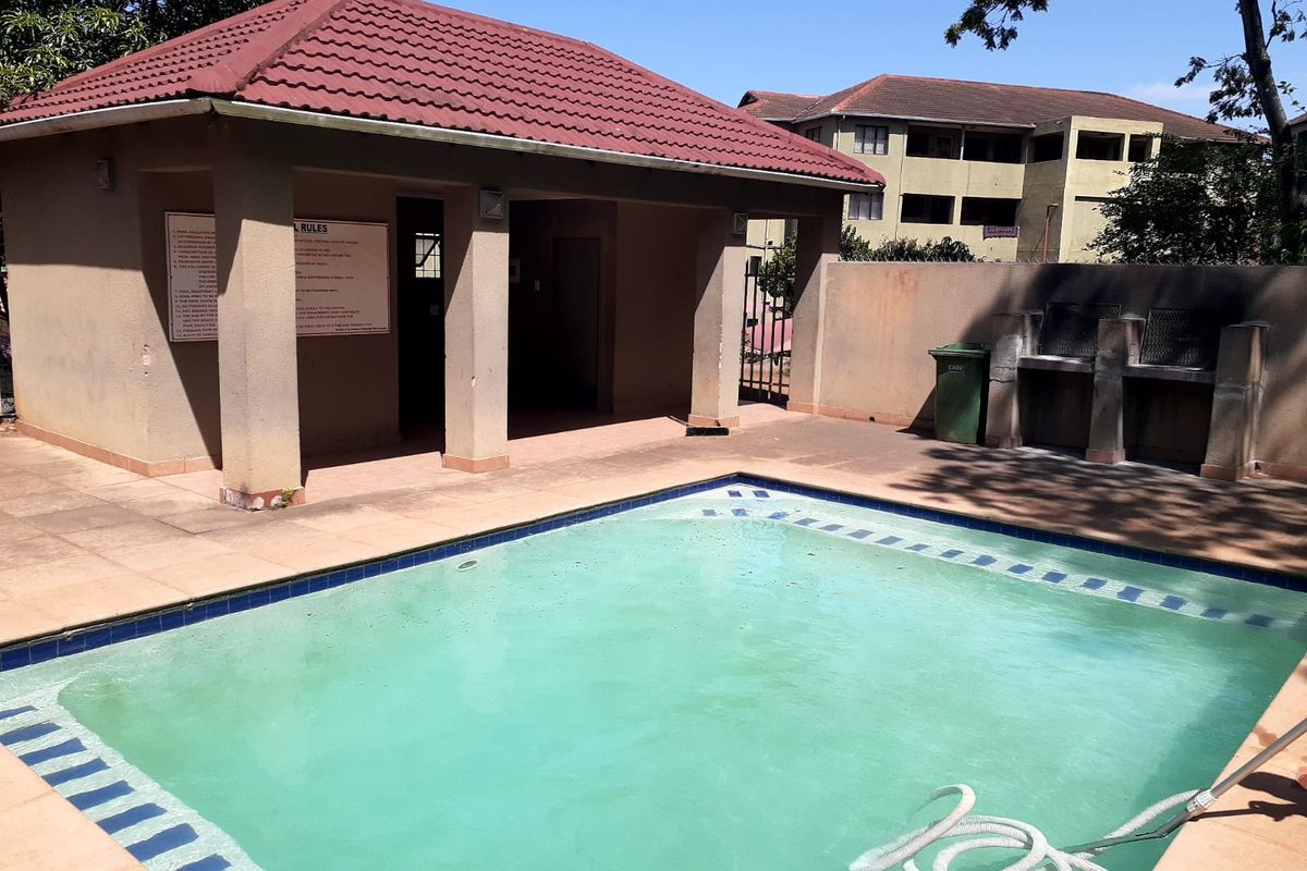 Communal swimming pool area featuring a covered patio structure, built-in braai facilities, and paved surround within an apartment complex.