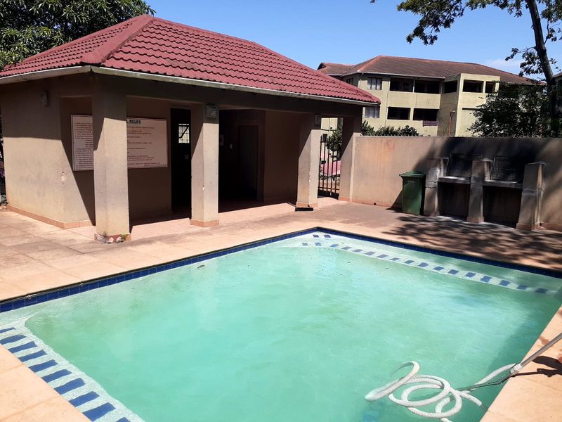Communal swimming pool area featuring a covered patio structure, built-in braai facilities, and paved surround within an apartment complex.