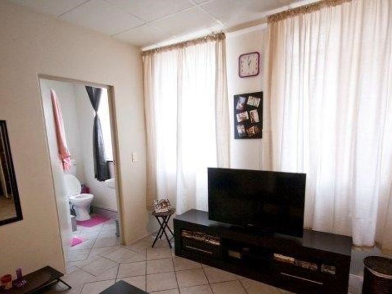 A living room area featuring tiled flooring and large windows with curtains, providing access to an adjacent bathroom.