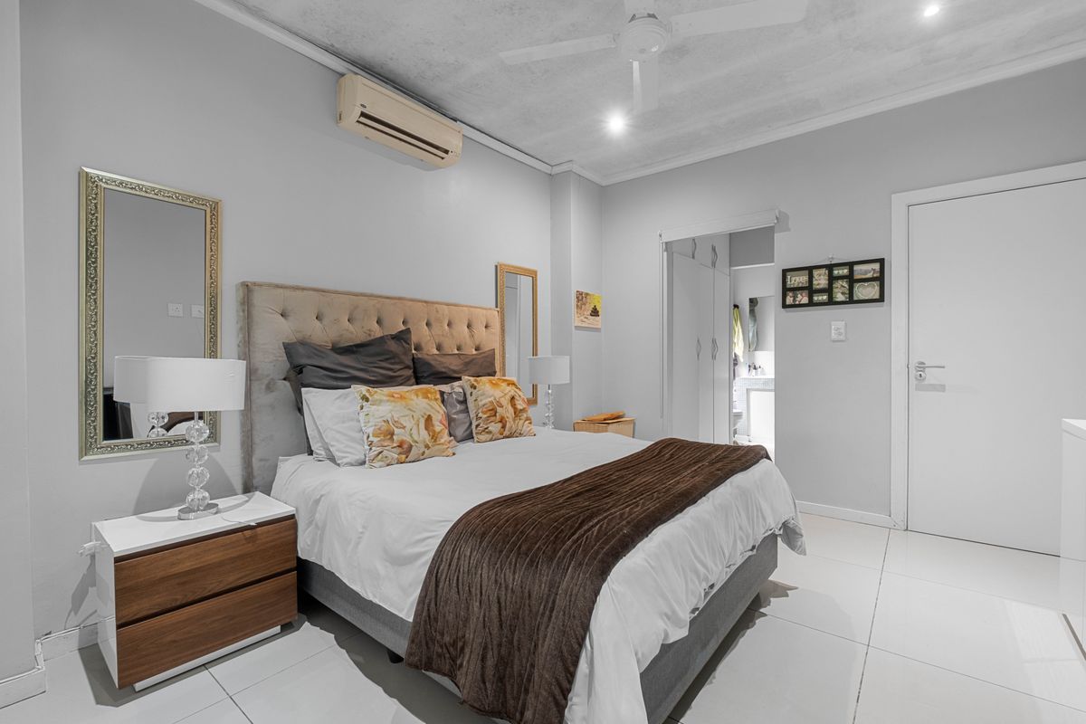 A well-lit bedroom featuring tiled flooring, a ceiling fan, and a wall-mounted air conditioning unit, with an adjoining en-suite bathroom.