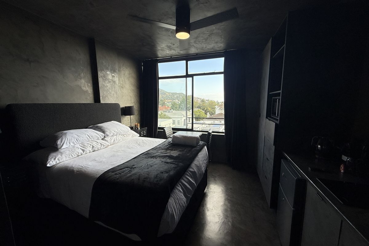 Modern bedroom featuring dark concrete-style walls, polished concrete flooring, a ceiling fan, and large windows offering scenic neighbourhood views.