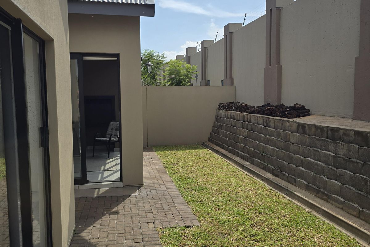 A private rear garden featuring a paved walkway, a small lawn area, and a retaining wall, enclosed by high perimeter walls with electric fencing.