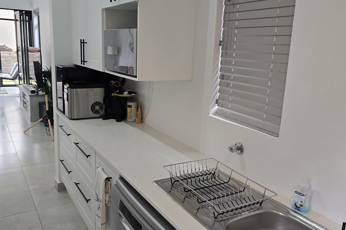 Modern kitchen featuring white cabinetry, stone countertops, integrated dishwasher, double stainless steel sink, and venetian blinds, with vaulted ceilings and tiled flooring.