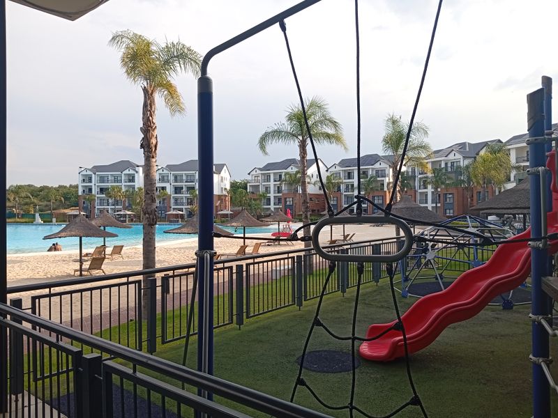 A view of the communal pool area featuring a sandy beach entry, thatched umbrellas, and a playground with fixed equipment.