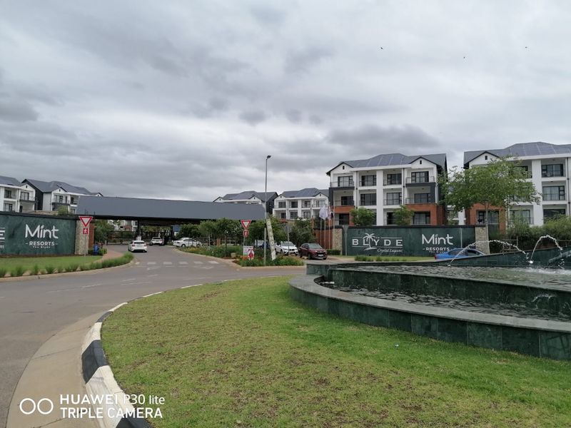 The entrance to The Blyde Mint Resorts featuring a landscaped roundabout with a water fountain, security gatehouse, and modern apartment buildings.