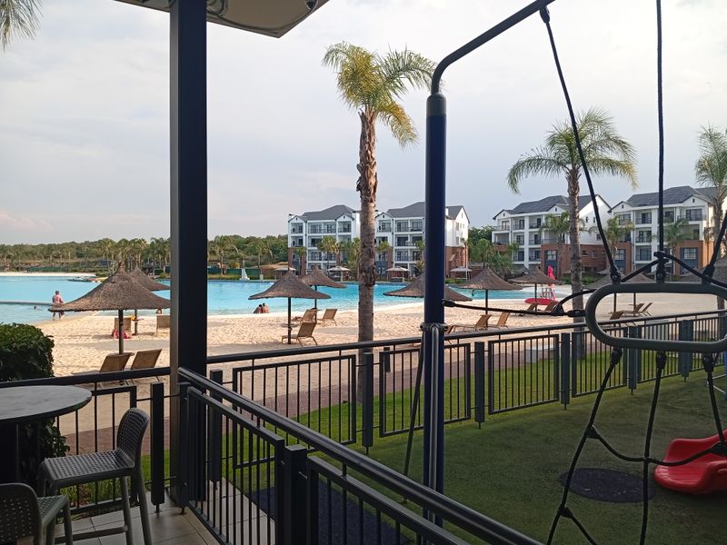 A covered patio area overlooking a communal beach-style swimming pool and landscaped grounds within a residential apartment complex.