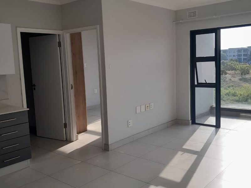 An open-plan living area featuring neutral tiled flooring, modern grey kitchen cabinetry, and a glass door leading to a balcony with natural light.