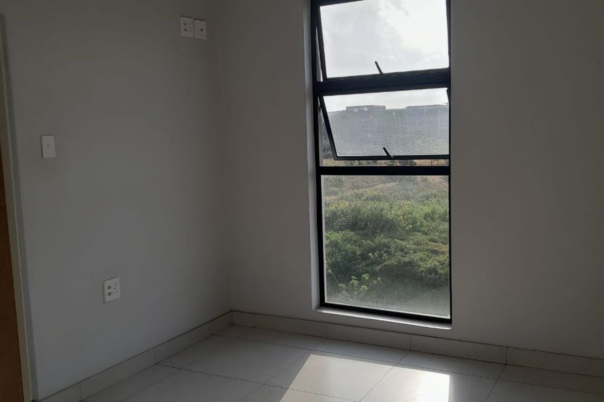 A bright, empty bedroom featuring neutral tiled flooring, a large vertical window with natural light, and a simple ceiling-mounted light fixture.