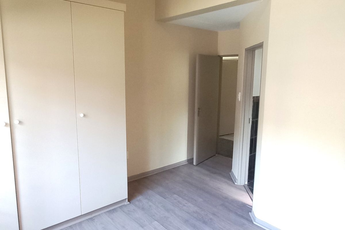A bright bedroom featuring built-in cupboards, vaulted ceilings, and modern wood-look laminate flooring, leading to an adjacent room.