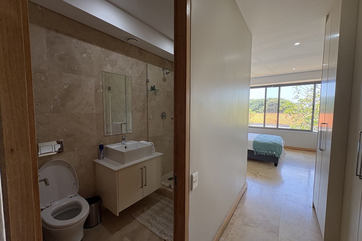 A modern en-suite bathroom featuring stone wall tiling, a floating vanity, and a glass-enclosed shower, adjacent to a bright bedroom with large windows.