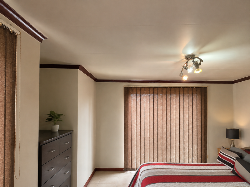 A bedroom featuring tiled flooring, dark wood-toned ceiling cornices, and vertical window blinds, providing a functional and neutral living space.