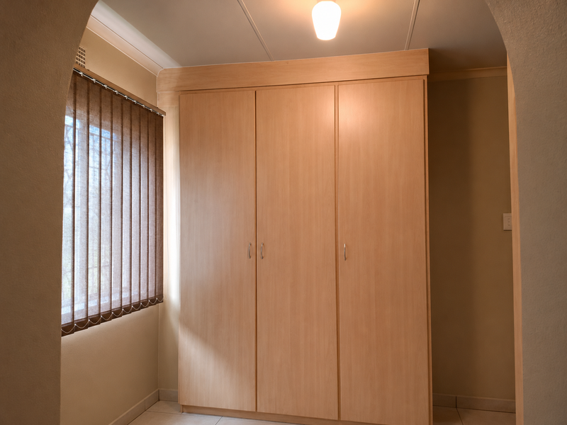 A clean bedroom space featuring light wood built-in cupboards, vertical window blinds, and a decorative archway entrance.