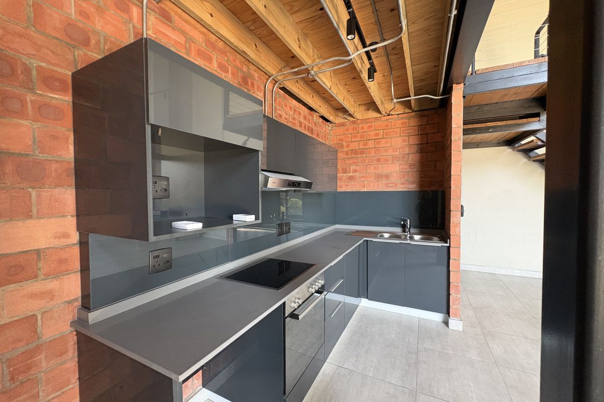 Modern industrial-style kitchen featuring exposed brick walls, timber ceiling beams, sleek grey cabinetry, integrated appliances, and a glass splashback.
