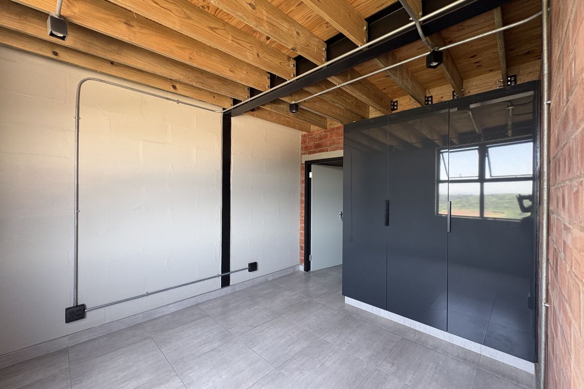 Modern industrial-style bedroom featuring exposed timber beams, brickwork, and sleek built-in cupboards with tiled flooring and exposed electrical conduit.