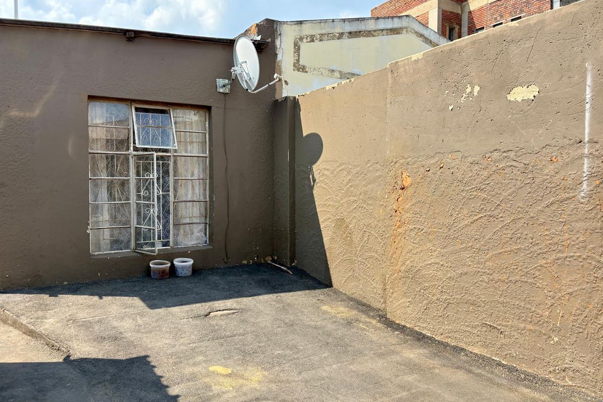 A small rear exterior courtyard area featuring a paved surface, a window with security bars, and a boundary wall requiring maintenance.