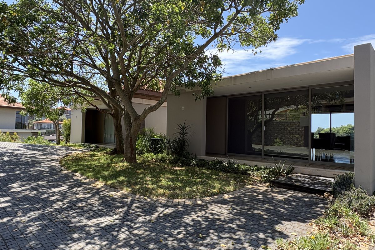 A modern single-story residence featuring large sliding glass doors, a paved driveway, and established trees in the front garden area.